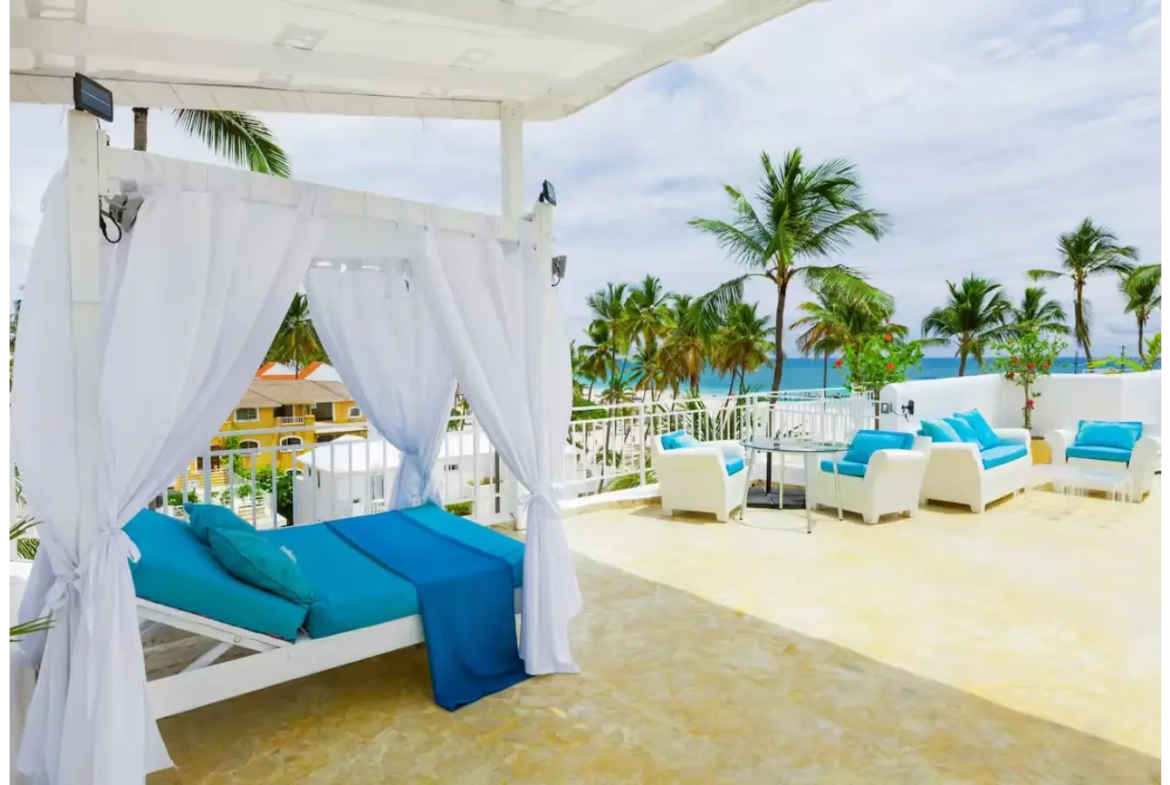 a white canopy with blue pillows and blue blankets on a patio with palm trees